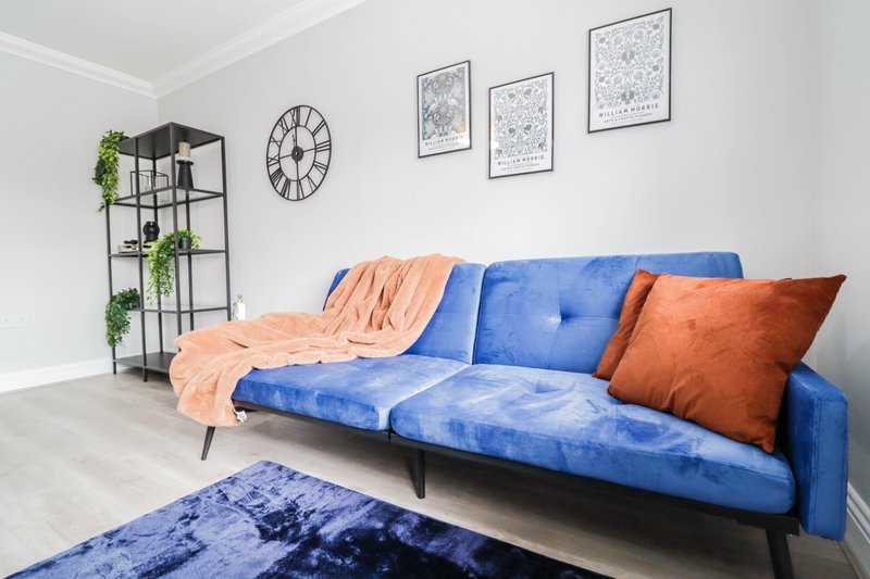 Modern living room with blue velvet sofa, orange cushions and throw, wall clock and framed prints