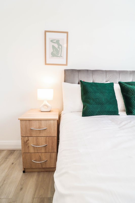 Bedroom with grey upholstered headboard, white bedding, green cushions, wooden nightstand and wall art