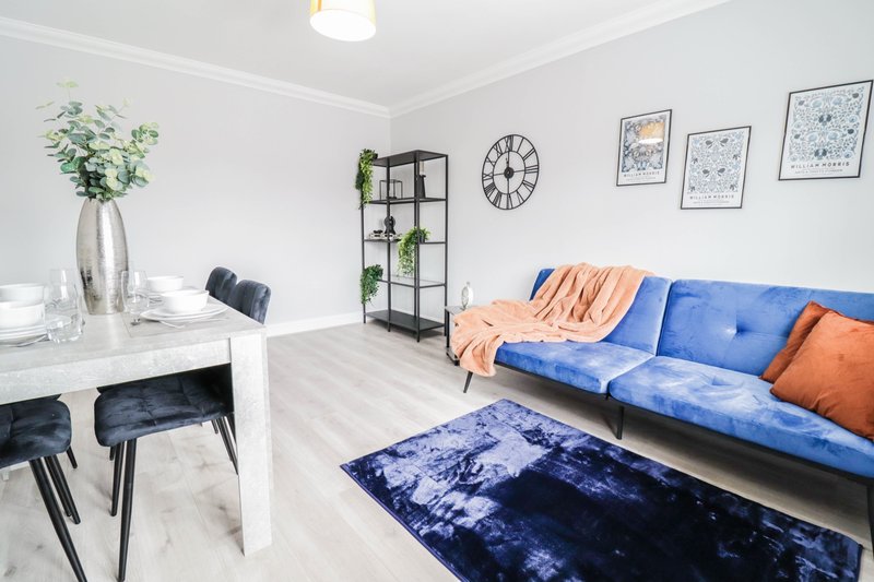 Open-plan living room with blue sofa, orange throw, dining table, potted plant and wall art