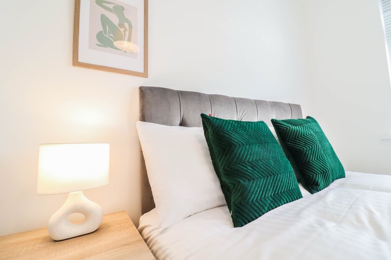 Modern bedroom with grey upholstered headboard, white bedding, green cushions and bedside lamp