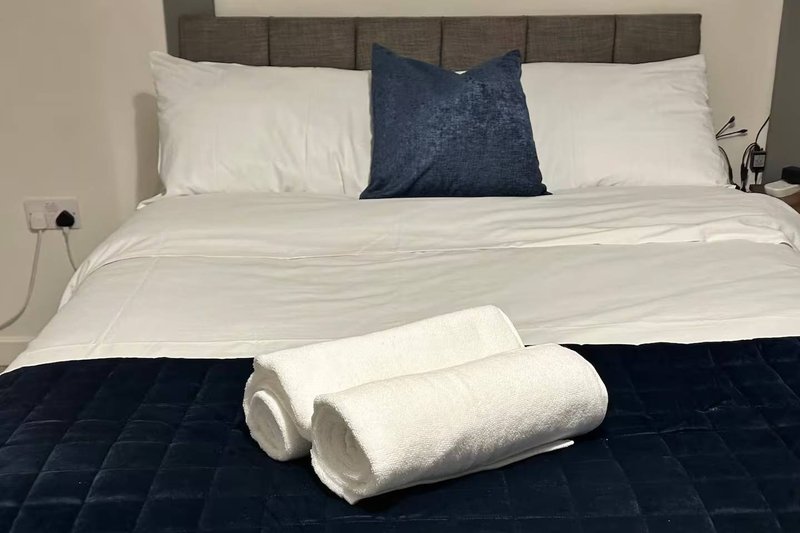 Bedroom with neatly made bed, white pillows, blue cushion, rolled towels and dark bedding