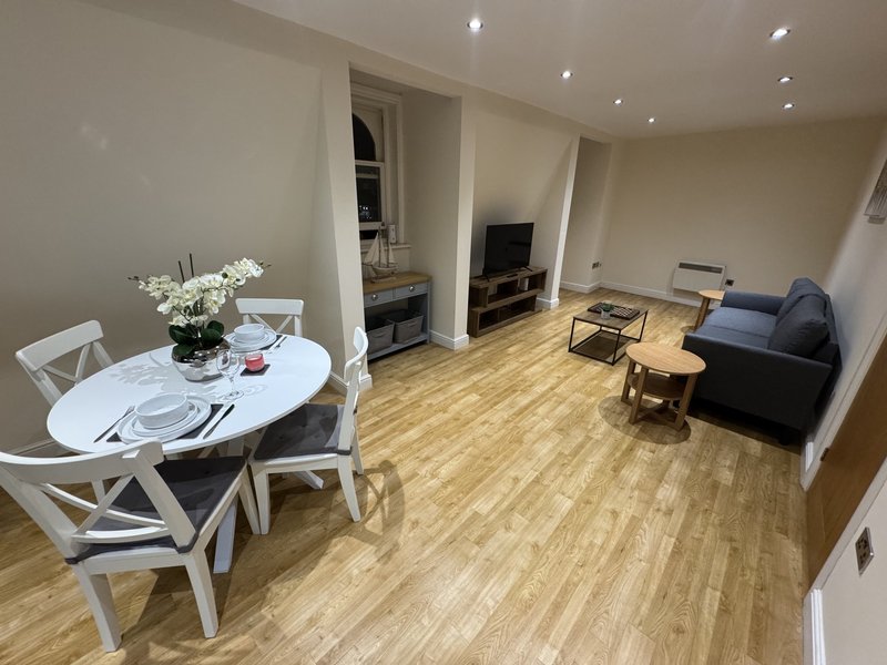 Open-plan living and dining area with white dining table, black sofa, TV, and wooden flooring