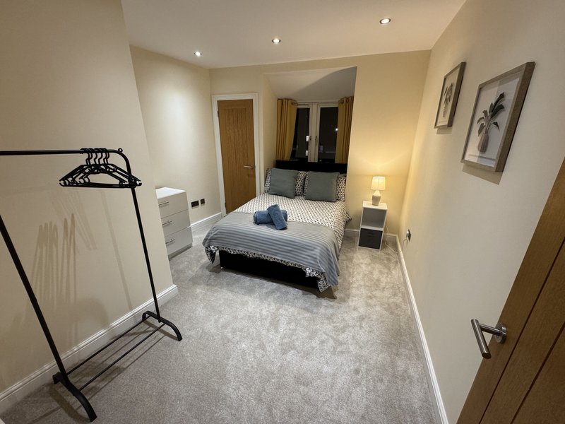 Modern bedroom with double bed, coat stand, ensuite bathroom visible, and neutral decor