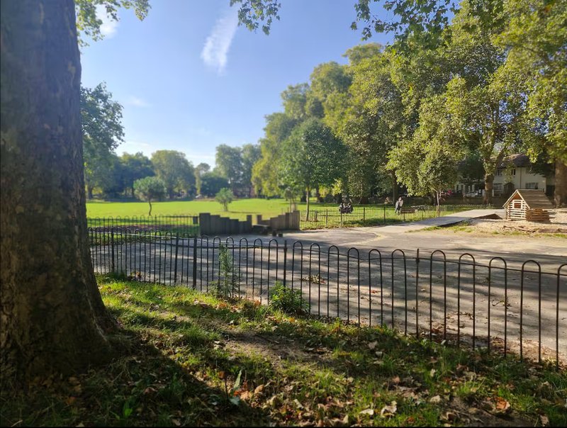 Tree-lined park with black metal fence, green grass, and paved pathway on sunny day