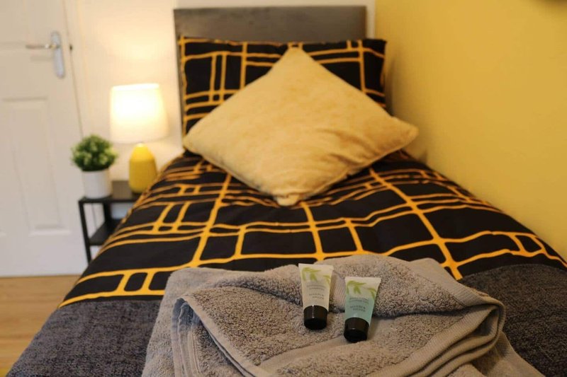 Bedroom with black and gold patterned bedding, yellow cushion, towels and toiletries on bed