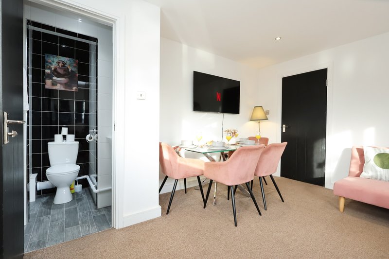 Modern open-plan living area with dining table, pink chairs, sofa, and en-suite bathroom visible