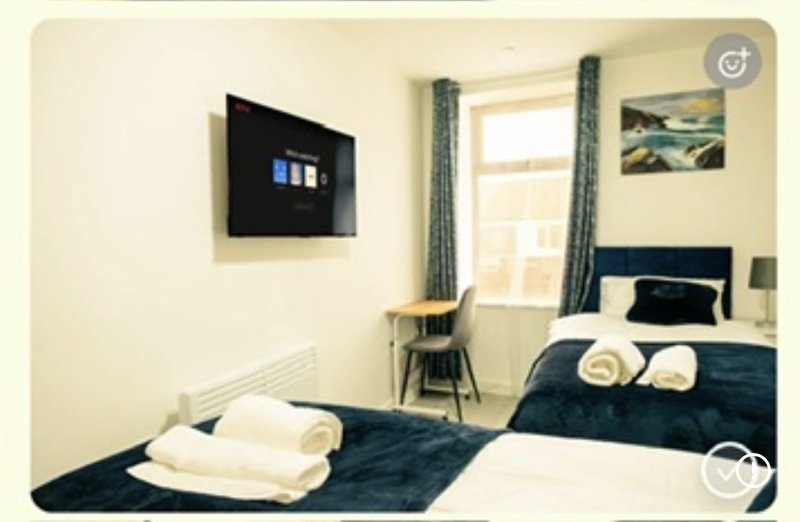 Bedroom with two single beds, dark bedding, window with curtains, wall-mounted TV, and wooden chair