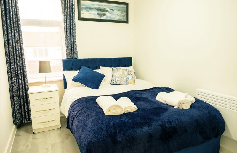 Bedroom with double bed, blue duvet, white pillows, cream bedside table and patterned curtains