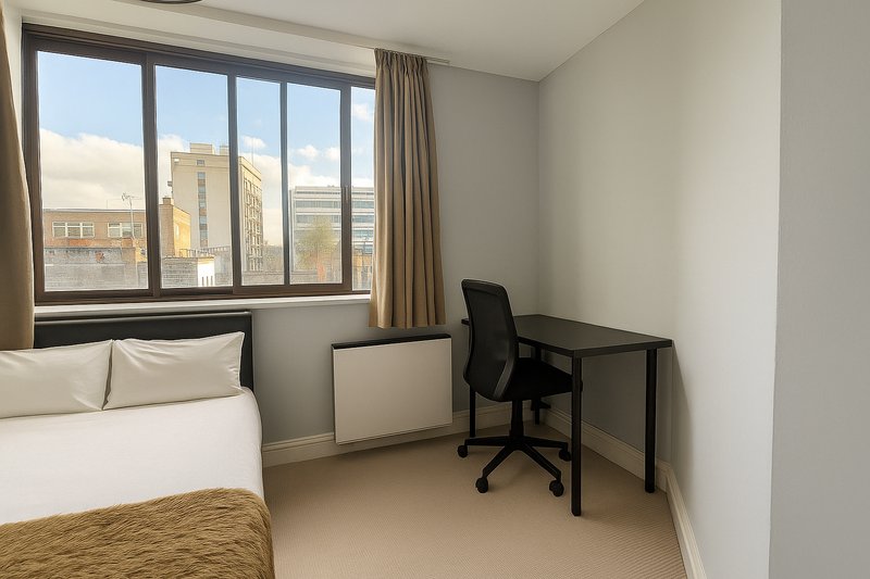 Bedroom with double bed, desk, office chair and large windows overlooking city