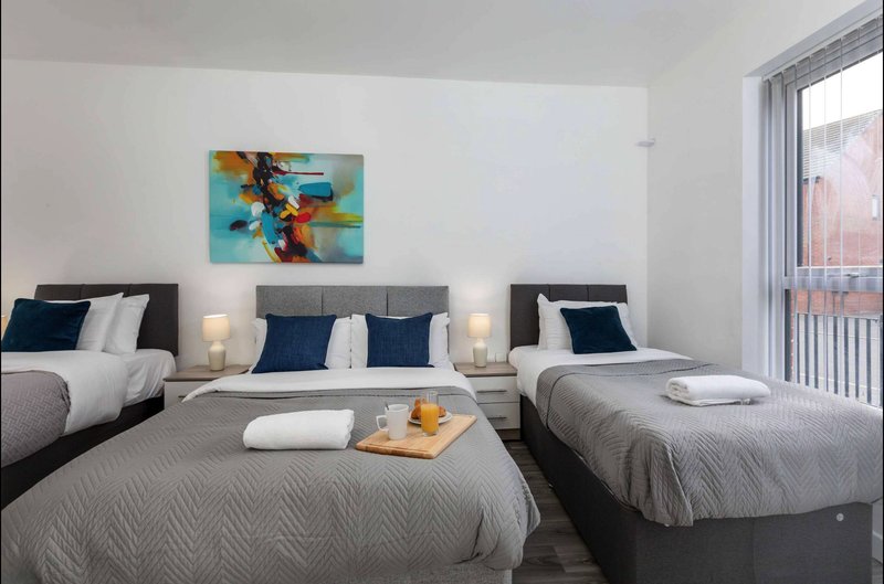 Modern bedroom with three single beds, white bedding, blue cushions, abstract wall art, and bright windows