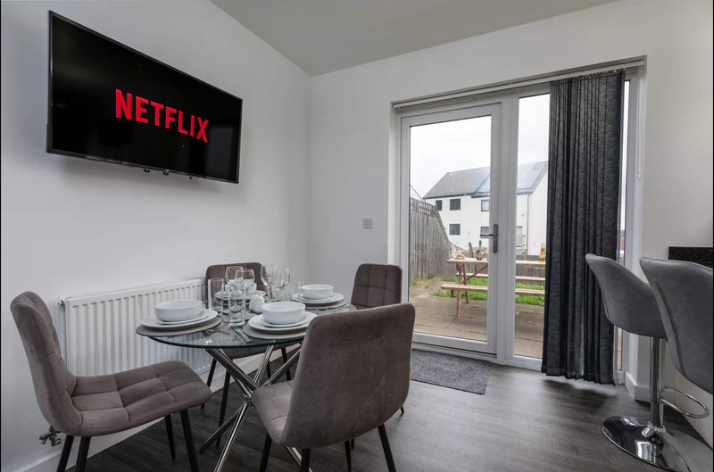 Open-plan living and dining area with wall-mounted TV, dining table, chairs, and patio doors overlooking garden
