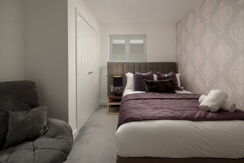 Bedroom with double bed, purple bedding, grey armchair, and patterned wallpaper