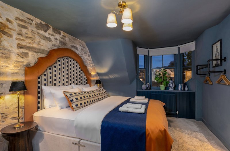 Modern bedroom with upholstered headboard, white bedding, blue and orange throw, and corner window with city views
