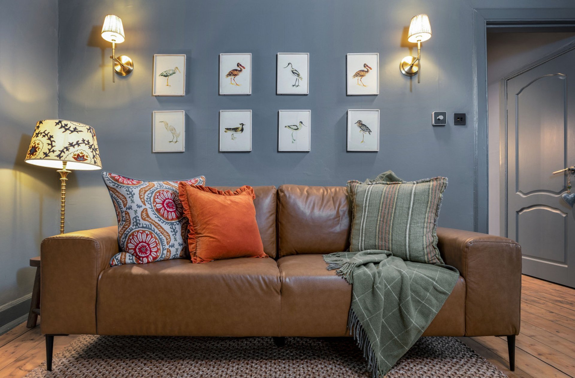 Living room with brown leather sofa, decorative pillows, throw blanket, wall art, and table lamp
