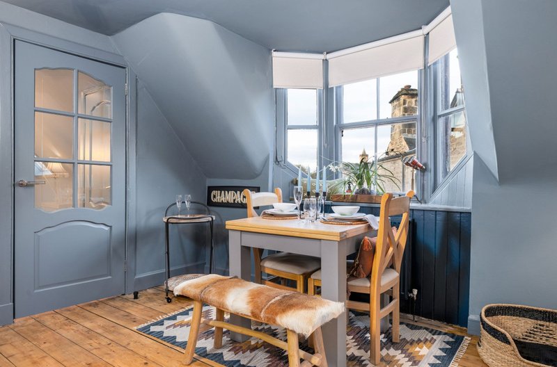 Bright attic dining room with wooden table, chairs, blue door and large windows overlooking street