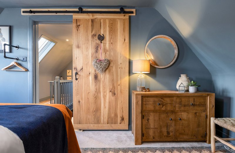 Modern bedroom with blue walls, wooden barn door, dresser, circular mirror and bed with blue bedding
