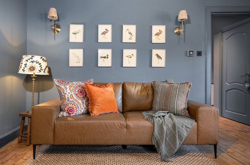 Living room with tan leather sofa, colorful cushions, wall art, and table lamp