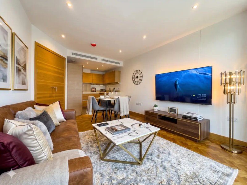 Modern living room with brown leather sofa, glass coffee table, wall-mounted TV, and open-plan kitchen area