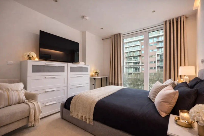 Modern bedroom with double bed, white dresser, wall-mounted TV, and city views through large windows