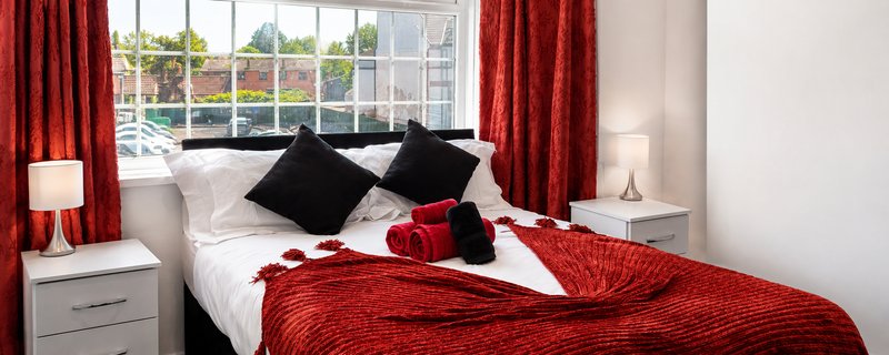 Modern bedroom with red and black bedding, white headboard, and large window with red curtains