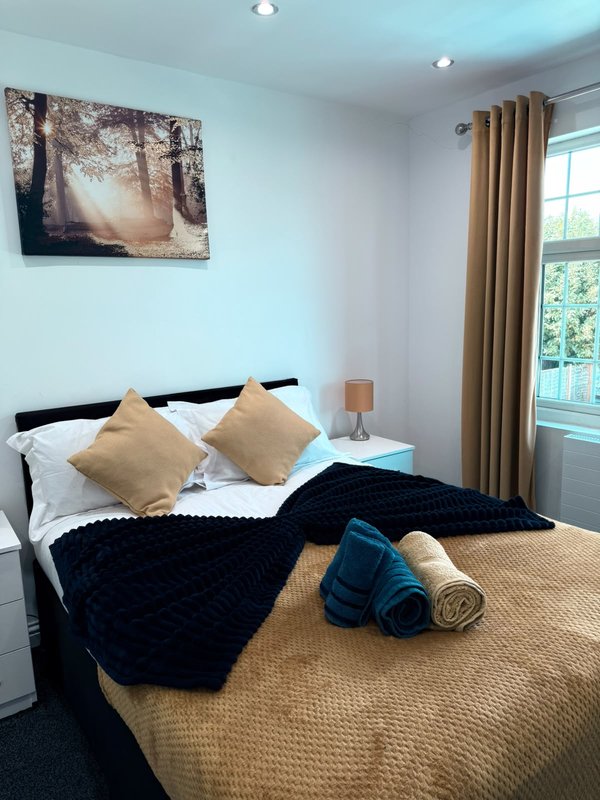 Bedroom with double bed, beige and black bedding, cushions, wall art, lamp and curtains