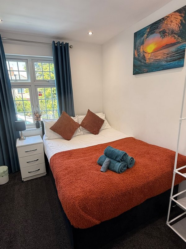 Bedroom with double bed, orange throw, brown cushions, window with blue curtains, artwork on wall