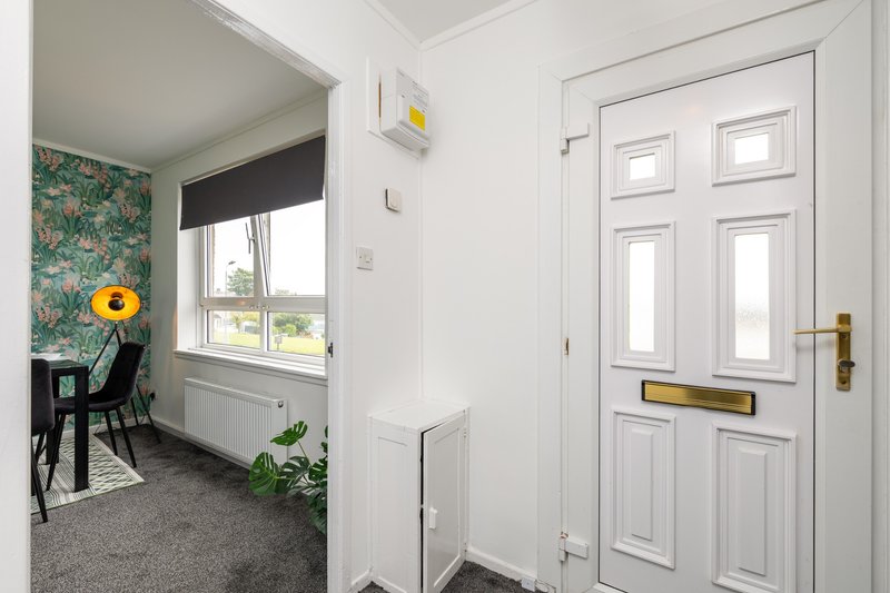 Bright hallway with white door, window with roller blind, and view into furnished room beyond