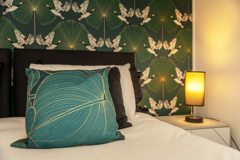 Modern bedroom with turquoise patterned cushion, white bedding, dark headboard, and decorative wallpaper with bird motifs