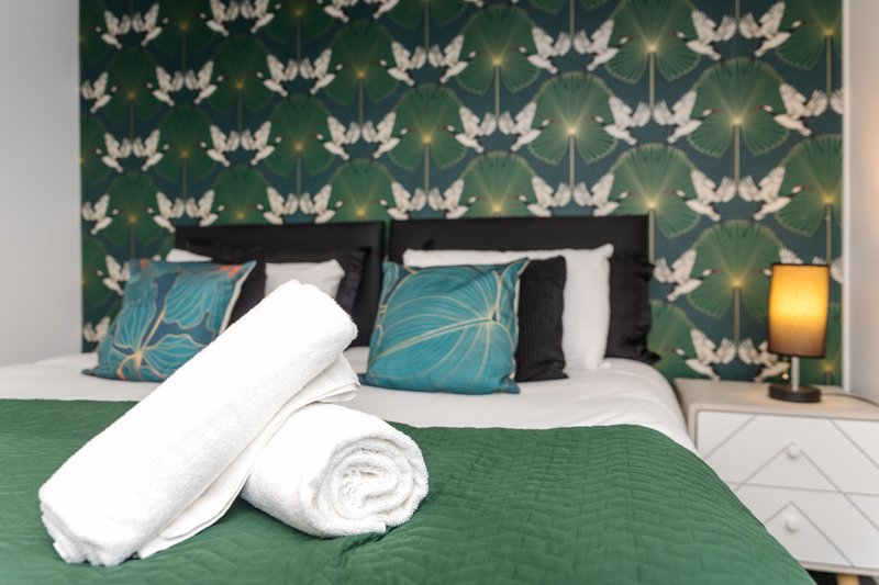 Bedroom with green patterned wallpaper, black bed frame, white pillows and rolled towels on green bedding