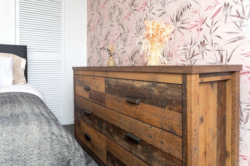Bedroom with wooden dresser, pink floral wallpaper, white louvered door, and bed with gray bedding