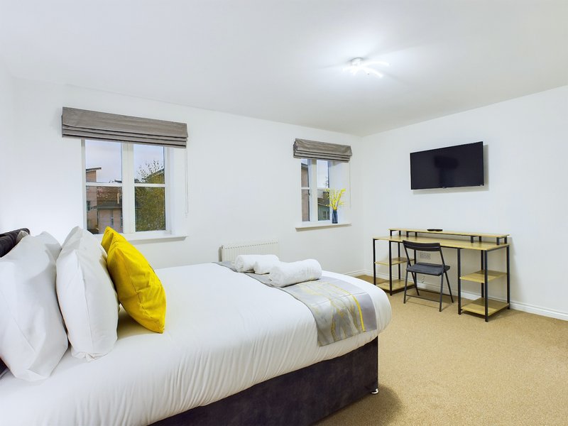 Bright bedroom with double bed, yellow cushion, wall-mounted TV and desk area