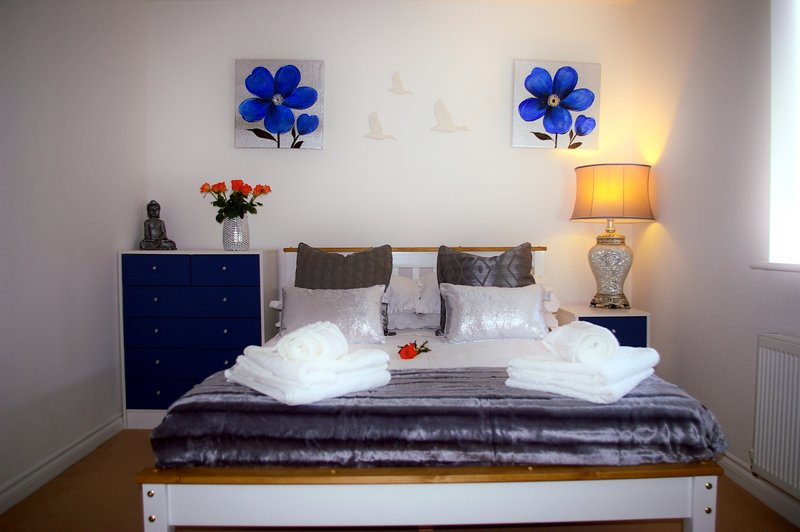 Bedroom with double bed, white pillows, dark bedspread, blue flower artwork, lamp and bedside table.