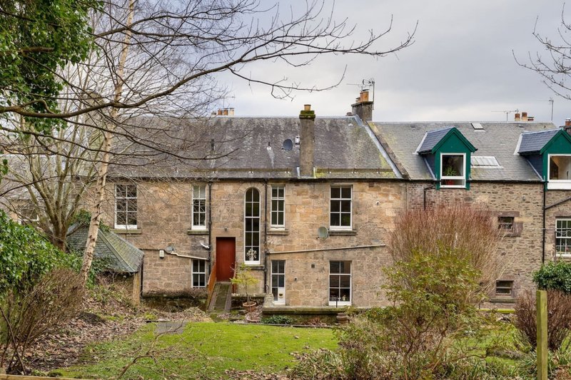 Stone Victorian building with multiple windows, red door and green roof dormers, surrounded by mature garden.
