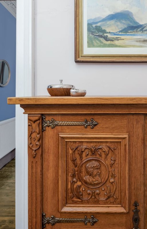 Wooden carved cabinet with decorative handles, framed landscape painting above, white wall