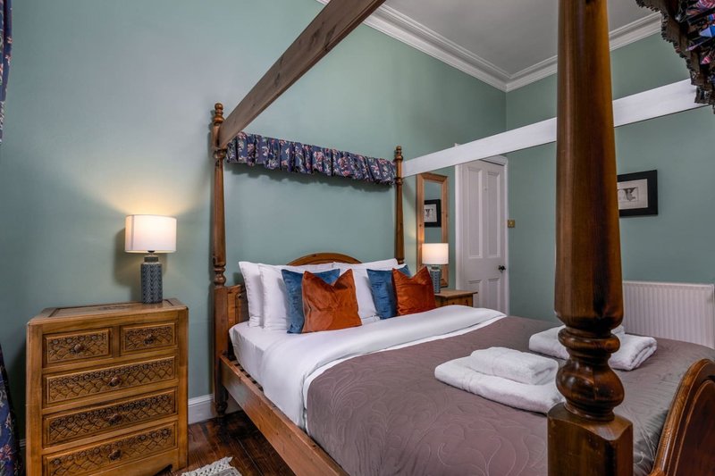 Bedroom with four-poster wooden bed, green walls, wooden dresser, and bedside lamp