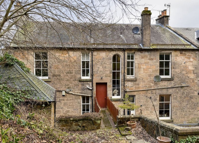 Stone cottage exterior with red door, multiple windows, chimneys and overgrown garden