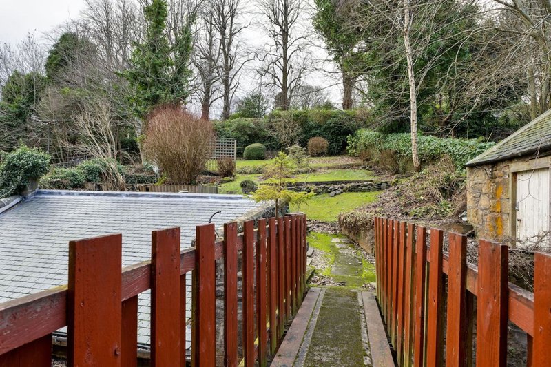 Garden pathway with wooden fencing, pond, trees and stone outbuilding in background
