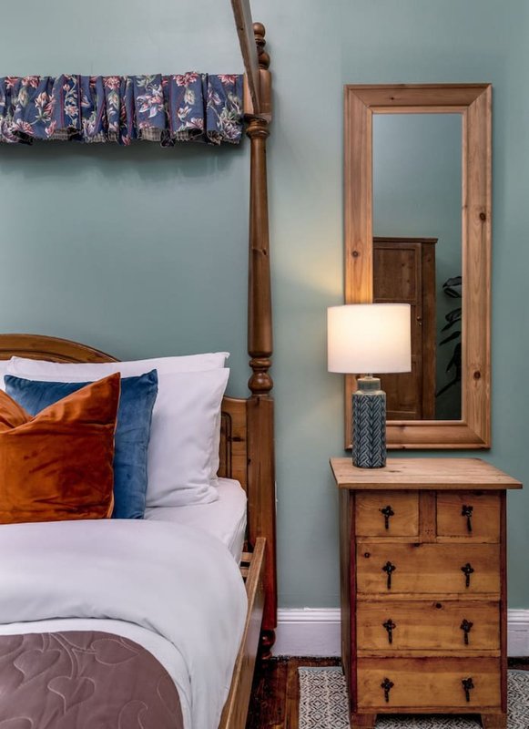 Bedroom with wooden four-poster bed, white linens, orange and blue pillows, bedside table with lamp and mirror