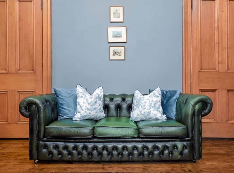 Green leather Chesterfield sofa with blue cushions and patterned pillows in blue-walled living room
