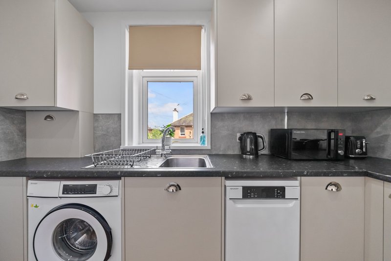 Modern kitchen with white cabinets, grey countertop, washing machine, dishwasher, and window overlooking garden
