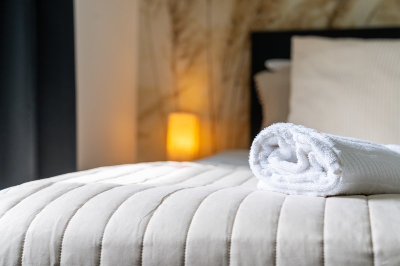 Rolled white towel on quilted bed with warm lighting and curtains