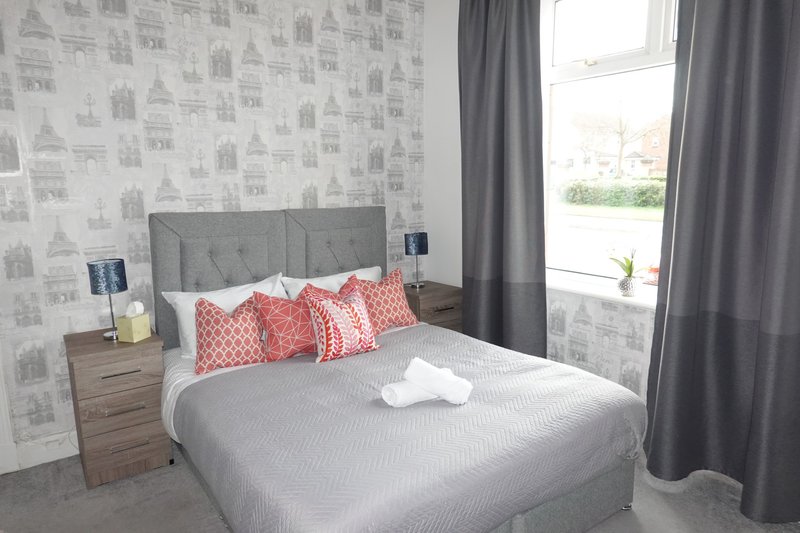 Bedroom with double bed, grey headboard, red patterned cushions, wooden nightstand and grey curtains