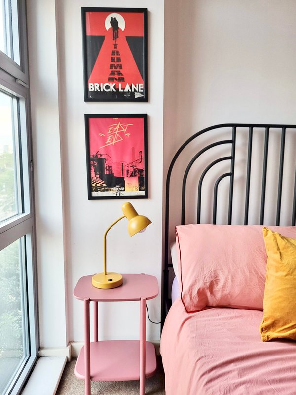 Bedroom corner with pink bed, metal headboard, side table, brass lamp, and wall art posters