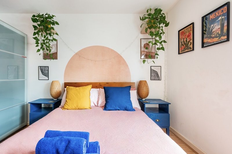 Modern bedroom with pink bedding, blue headboard, yellow and blue cushions, potted plants and wall art