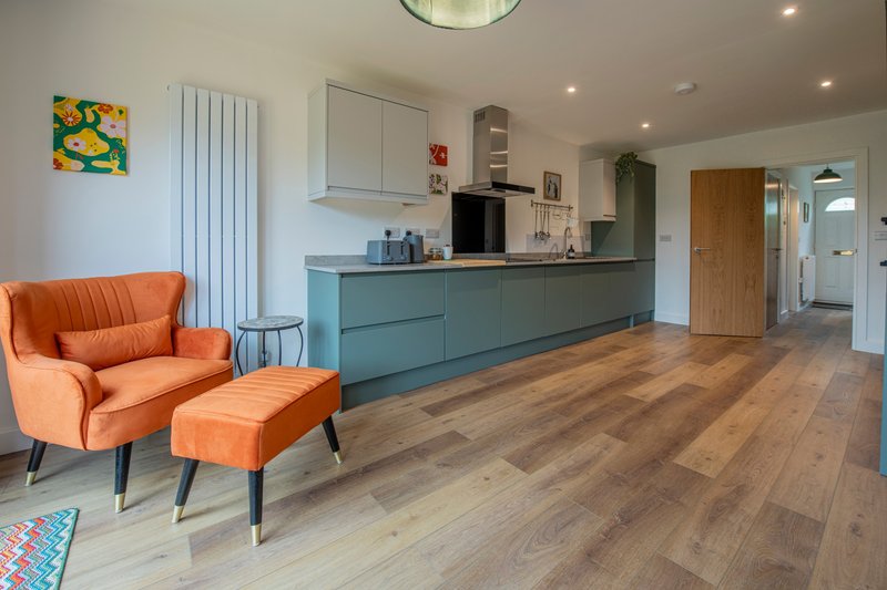 Modern open-plan kitchen and lounge with orange armchair, teal cabinetry and wooden flooring