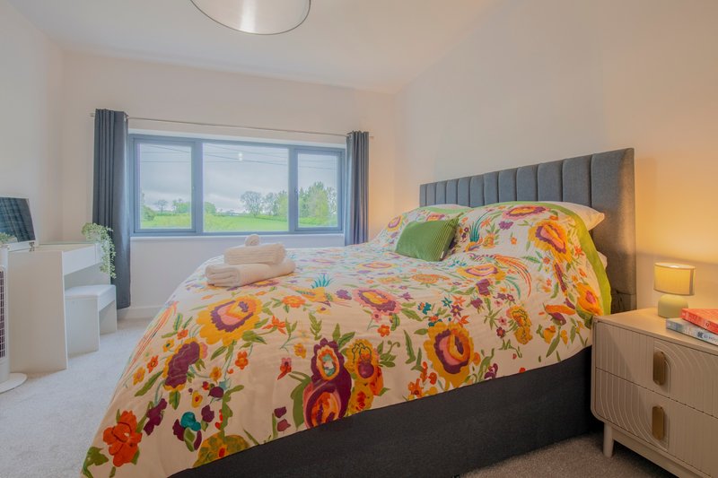 Bright bedroom with double bed, colorful floral bedding, grey headboard, wooden nightstands and large window