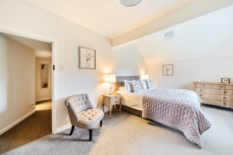 Bright bedroom with double bed, upholstered chair, wooden dresser and ensuite entrance