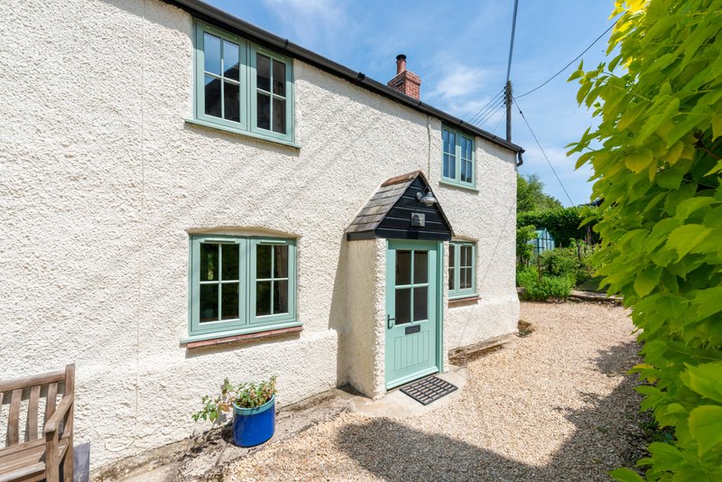 White rendered cottage exterior with green door, windows, gravel driveway and garden