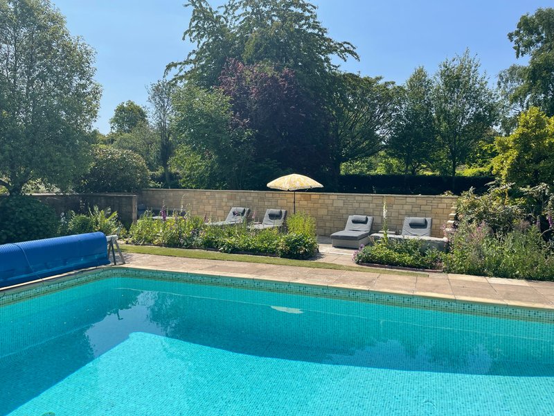 Swimming pool with blue water, stone patio, garden seating areas and trees