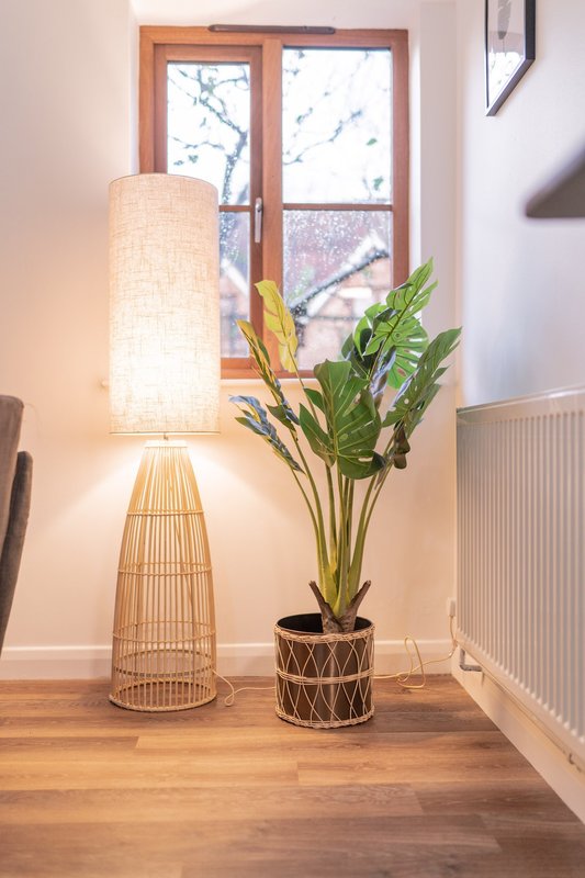 Bright interior corner with wooden floor, potted plant, woven floor lamp, and window overlooking trees